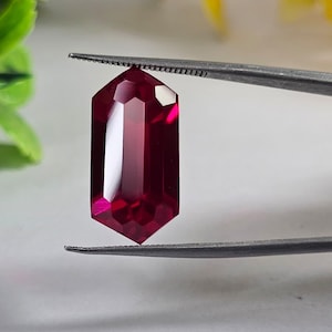 May include: A faceted, elongated ruby gemstone held by tweezers. The gem is a deep, rich red color with multiple reflective surfaces. The shape is a modified hexagon, showcasing the stone's clarity and cut.