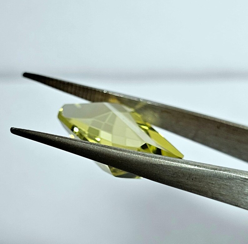 Pode incluir: Uma gema facetada, amarelo claro, &eacute; segurada por uma pin&ccedil;a prateada contra um fundo branco. A gema tem formato de p&ecirc;ra com m&uacute;ltiplas facetas que refletem a luz. A pin&ccedil;a est&aacute; em foco, destacando a clareza da gema.