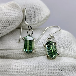 May include: A pair of silver earrings featuring green, rectangular gemstones. The gemstones are faceted and have an emerald-like hue. The earrings are presented on a white, textured surface.