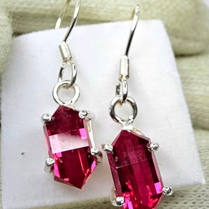 May include: A pair of dangle earrings with faceted, elongated, ruby-red gemstones. The gemstones are set in silver-coloured metal with small, round accents. The earrings have a simple hook design.