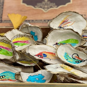 May include: A collection of oyster shells painted with colorful fishing lures. The shells are arranged in a wooden box, with some of the shells overlapping.