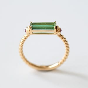 May include: A gold ring featuring a rectangular green gemstone and two small red gemstones. The ring has a twisted band design. The gemstones are set in a gold setting. The ring is displayed on a white background.