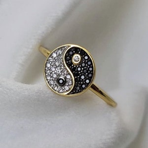 May include: A gold-toned ring featuring a yin-yang design. The yin side is black, studded with small black stones, while the yang side is white, covered in small white stones. A small black stone and a white stone are at the center of each side.