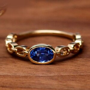 May include: A gold ring featuring a blue oval sapphire gemstone set in a bezel setting. The band is crafted with a chain-like design and small round gemstones.