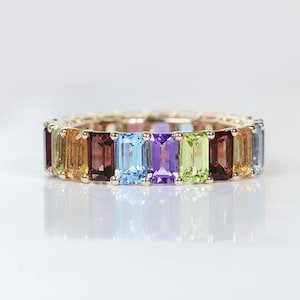 May include: A gold band ring featuring a row of colourful gemstones. The gemstones are emerald cut and include amethyst, citrine, peridot, topaz, and garnet.