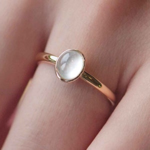 May include: A delicate gold ring featuring a white oval gemstone. The ring is simple and elegant.