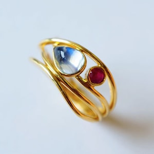 May include: A gold ring with three bands, featuring a teardrop-shaped moonstone and a round ruby.