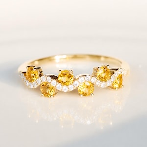 May include: A gold ring featuring a wavy band design set with five round yellow gemstones and a row of small white accent stones. The band has a polished finish and follows a curved, undulating pattern across the top of the ring setting.