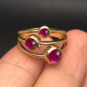 May include: Gold ring with three red gemstones. The ring is made of three bands that are intertwined.