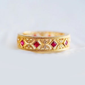 May include: A gold band ring with a delicate filigree design featuring red gemstones and smaller clear gemstones.