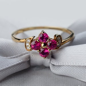 Puede incluir: Un anillo de oro con un grupo de cinco piedras preciosas rosas engastadas en forma de flor. El anillo está adornado con pequeñas piedras preciosas transparentes.