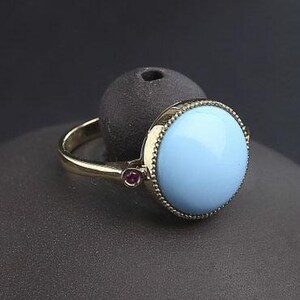 May include: A gold ring with a light blue cabochon gemstone set in a bezel setting. The ring has two small ruby accents on either side of the main stone.