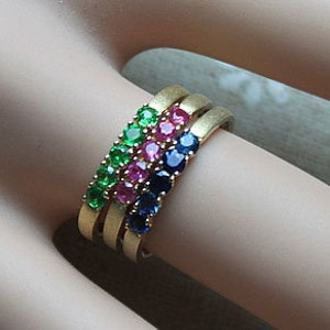 Op de afbeelding: Drie gouden ringen met een rij edelstenen. De bovenste ring heeft groene edelstenen, de middelste ring heeft roze edelstenen en de onderste ring heeft blauwe edelstenen.
