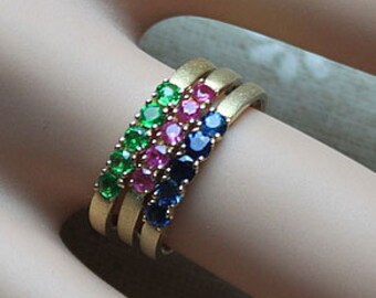 Op de afbeelding: Drie gouden ringen met een rij edelstenen. De bovenste ring heeft groene edelstenen, de middelste ring heeft roze edelstenen en de onderste ring heeft blauwe edelstenen.
