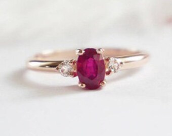 May include: A rose gold engagement ring featuring an oval ruby gemstone with two smaller diamond accents.