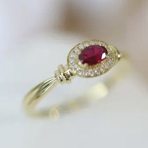 Puede incluir: Un anillo de oro con una piedra preciosa ovalada de rubí rojo rodeada por un halo de diamantes más pequeños.