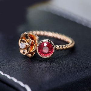 May include: A rose gold ring with a red gemstone and a smaller diamond set in a rose-shaped design. The band is twisted.