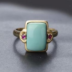 Może przedstawiać: Złoty pierścionek z prostokątnym turkusowym kamieniem i dwoma małymi rubinowymi kamieniami po bokach.