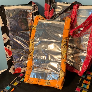 May include: Three insulated lunch bags with different patterns. The bags are made of silver material with a clear plastic window. The bags have black, red, and orange fabric trim.