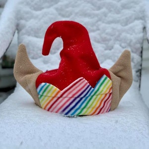Op de afbeelding: Een rode en witte elfenmuts met regenboogstrepen en bruine oorflappen. De muts ligt in de sneeuw.