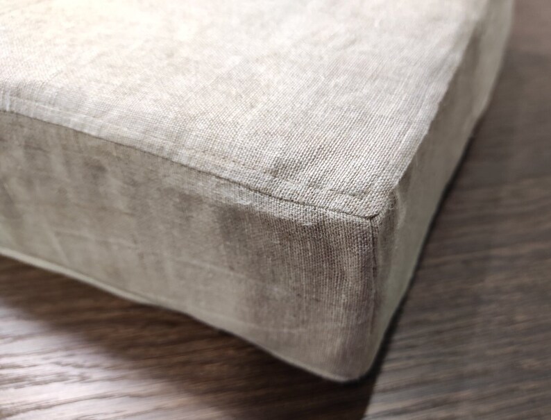 Puede incluir: Primer plano de un coj&iacute;n de lino beige con una costura bien cosida. El coj&iacute;n est&aacute; sobre una superficie de madera.