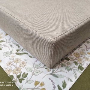 May include: A beige square cushion with a textured fabric cover. The cushion is sitting on a white fabric with a floral pattern.