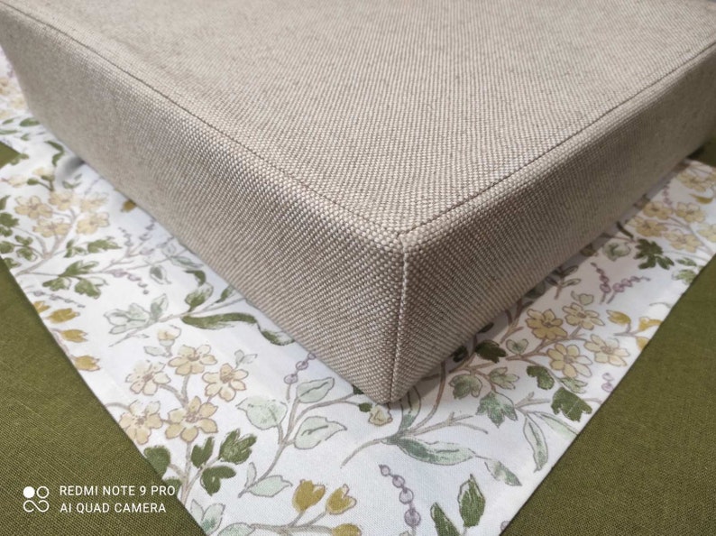 May include: A beige square cushion with a textured fabric cover. The cushion is sitting on a white fabric with a floral pattern.