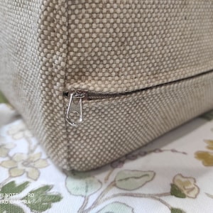 May include: Close-up of a beige fabric cushion with a zipper closure. The zipper is silver and has a small metal pull tab.
