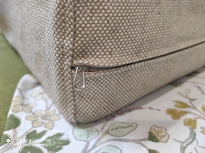 May include: Close-up of a beige fabric cushion with a zipper closure. The zipper is silver and has a small metal pull tab.