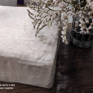 May include: A square, beige linen cushion with a subtle texture. The cushion is sitting on a dark wood surface.