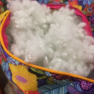 K&ouml;nnte beinhalten: Eine bunte Stofftasche mit Rei&szlig;verschluss&ouml;ffnung, gef&uuml;llt mit wei&szlig;er F&uuml;llung. Die Tasche hat ein Blumenmuster mit gelben, blauen, rosa und gr&uuml;nen Blumen.