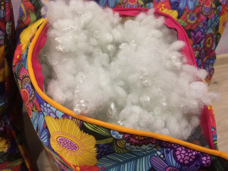 K&ouml;nnte beinhalten: Eine bunte Stofftasche mit Rei&szlig;verschluss&ouml;ffnung, gef&uuml;llt mit wei&szlig;er F&uuml;llung. Die Tasche hat ein Blumenmuster mit gelben, blauen, rosa und gr&uuml;nen Blumen.