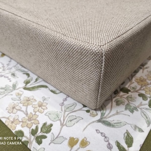 May include: A beige fabric cushion with a square shape and a subtle texture. The cushion is sitting on a white fabric with a floral pattern.