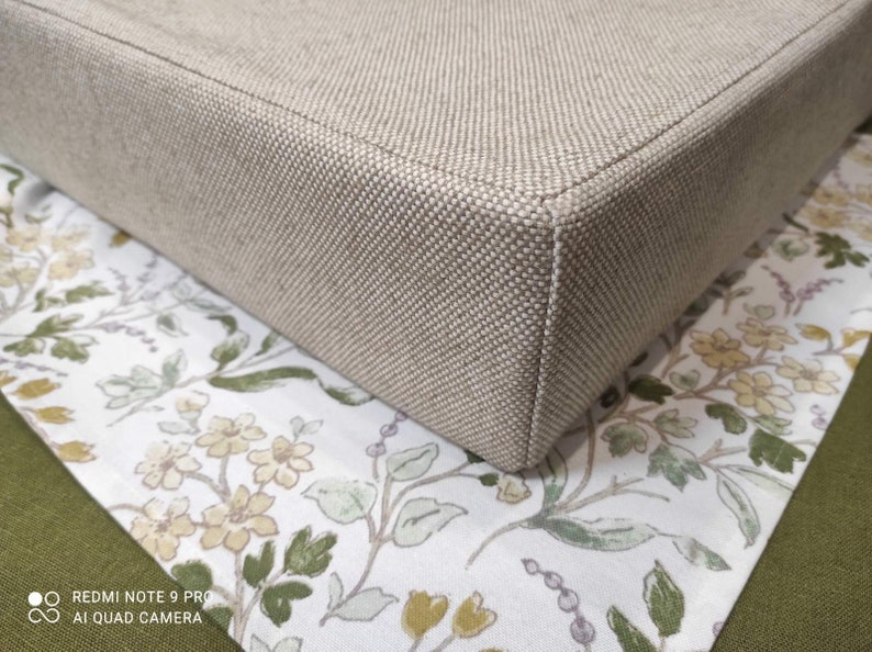 May include: A beige fabric cushion with a square shape and a subtle texture. The cushion is sitting on a white fabric with a floral pattern.