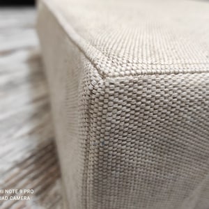 Peut inclure: Gros plan d'un ottoman en tissu beige avec une texture tissée. L'ottoman a une forme carrée et une surface lisse et uniforme.