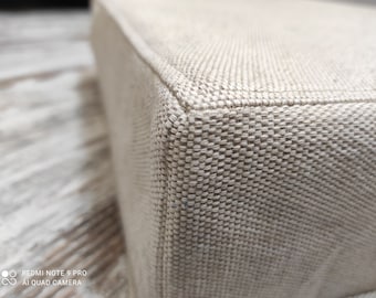 Puede incluir: Primer plano de un puf de tela beige con textura tejida. El puf tiene forma cuadrada y una superficie lisa y uniforme.