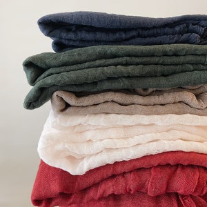 May include: A stack of six folded, lightweight fabric cloths in various colors. The colors include navy blue, dark green, tan, off-white, and red. The cloths have a slightly textured appearance and are neatly arranged.