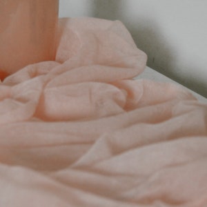May include: A soft, light peach-colored fabric is draped across a surface, with a cylindrical object partially visible in the background. The fabric is gathered and folded, creating a textured effect. The overall aesthetic is delicate and airy.
