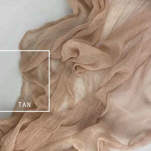 May include: A close-up of a tan sheer fabric with a soft, flowing texture. The fabric is draped and folded, creating a sense of movement and lightness. The word "TAN" is written in white text on the fabric.