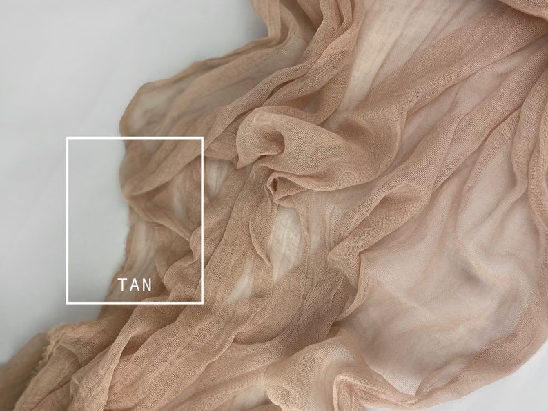 May include: A close-up of a tan sheer fabric with a soft, flowing texture. The fabric is draped and folded, creating a sense of movement and lightness. The word "TAN" is written in white text on the fabric.