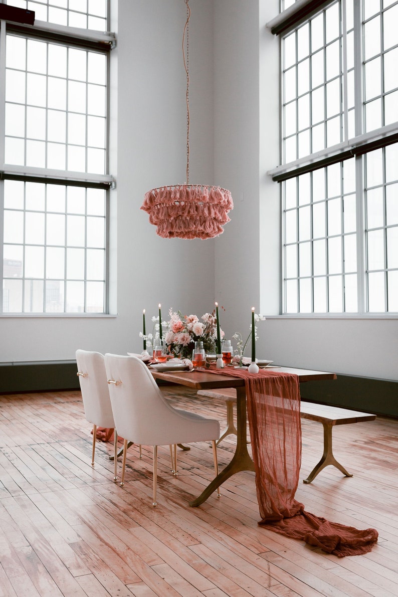 K&ouml;nnte beinhalten: Ein langer Holztisch mit einem rosafarbenen Quasten-Kronleuchter, der dar&uuml;ber h&auml;ngt. Der Tisch ist mit wei&szlig;en St&uuml;hlen, einem rosafarbenen Blumenarrangement und einem langen, rostfarbenen L&auml;ufer gedeckt.