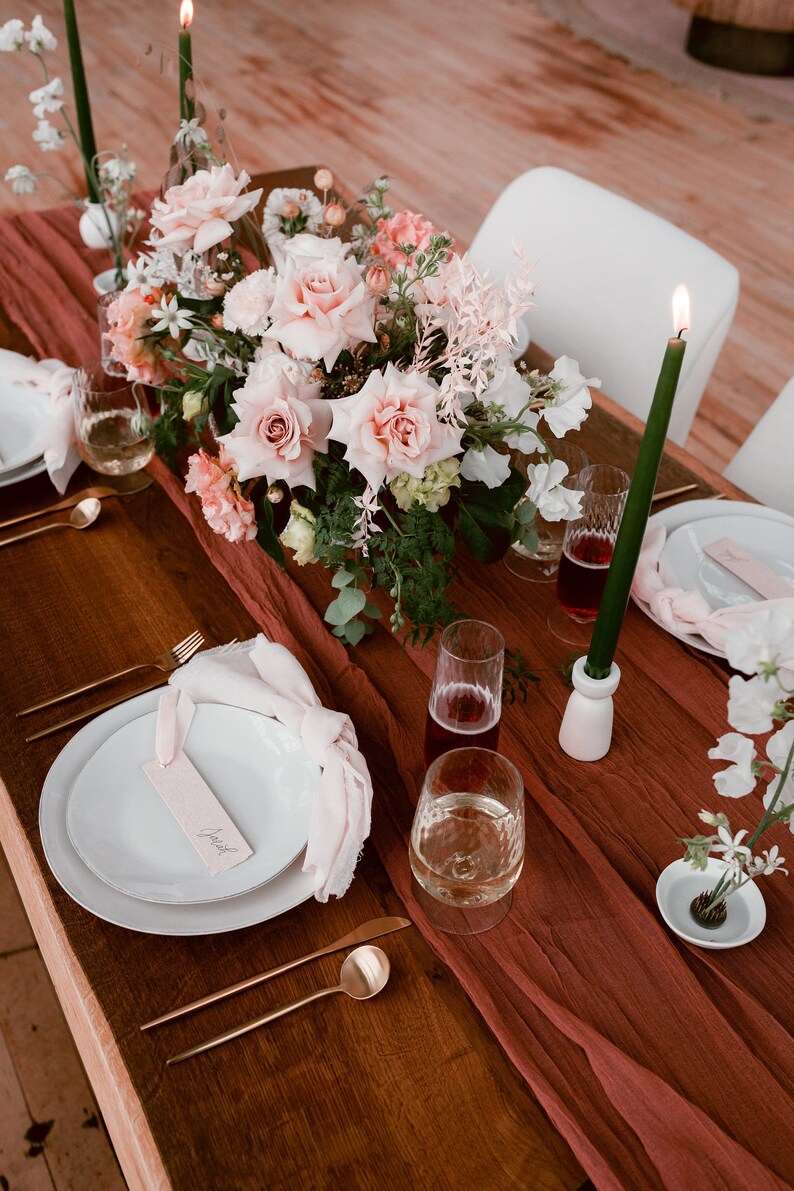 K&ouml;nnte beinhalten: Ein rustikaler Holztisch mit einem Mittelst&uuml;ck aus rosa Rosen und Gr&uuml;npflanzen. Der Tisch ist mit einem burgunderfarbenen Leinenl&auml;ufer bedeckt und verf&uuml;gt &uuml;ber wei&szlig;e Teller, goldenes Besteck und Gl&auml;ser, die mit Wein und Wasser gef&uuml;llt sind. Der Platzkarte steht "Josh" geschrieben.
