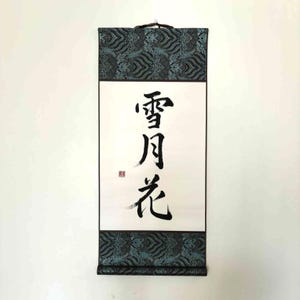 Könnte beinhalten: Eine traditionelle japanische Schriftrolle mit schwarzer Kalligraphie auf weißem Papier. Die oberen und unteren Ränder weisen einen teal-schwarzen Stoff mit Muster auf. Die Schriftrolle hängt an einer weißen Wand.