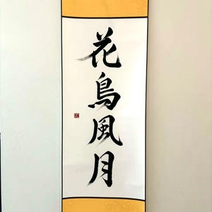 May include: A vertical scroll featuring Japanese calligraphy in black ink on white paper, with gold borders at the top and bottom. The calligraphy reads "Flower, Bird, Wind, Moon". A small red seal is present.