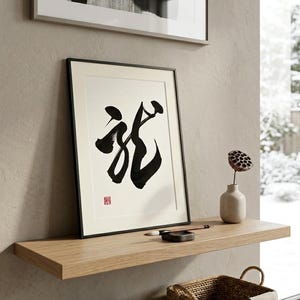 Könnte beinhalten: Gerahmtes Kunstwerk mit kräftiger schwarzer Kalligraphie auf weißem Hintergrund, vor einer neutralen Wand. Darüber ein kleinerer schwarz-weißer abstrakter Druck. Ein Holzregal zeigt eine Vase, einen Pinsel und einen Korb.