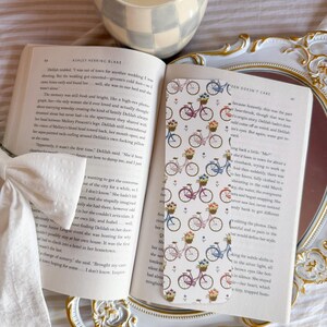 May include: A bookmark with a bicycle pattern, featuring bicycles in blue, pink, and purple with flower baskets. The bookmark is placed in an open book with text, next to a decorative candle and a mirror.