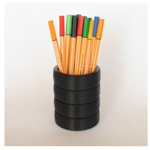 May include: A black tire-shaped pencil holder with a stack of colorful pens inside. The holder is made of recycled tire rubber.