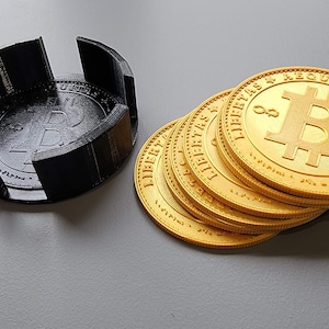 May include: A stack of six gold-colored Bitcoin coins with the inscription "LIBERTAS ÆQUITATAS VERITAS" around the edge. The coins are sitting on a gray surface next to a black 3D printed coin holder.