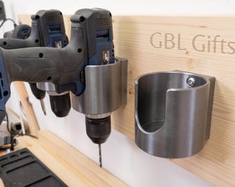 May include: A light-colored wooden wall-mounted rack featuring the text GBL Gifts, equipped with two grey metal cylindrical holders. One holder supports a blue and black cordless power drill, while the other holder remains empty and ready for use.