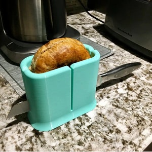 May include: A teal bagel slicer with a bagel inside and a silver knife attached. The slicer is on a speckled countertop.